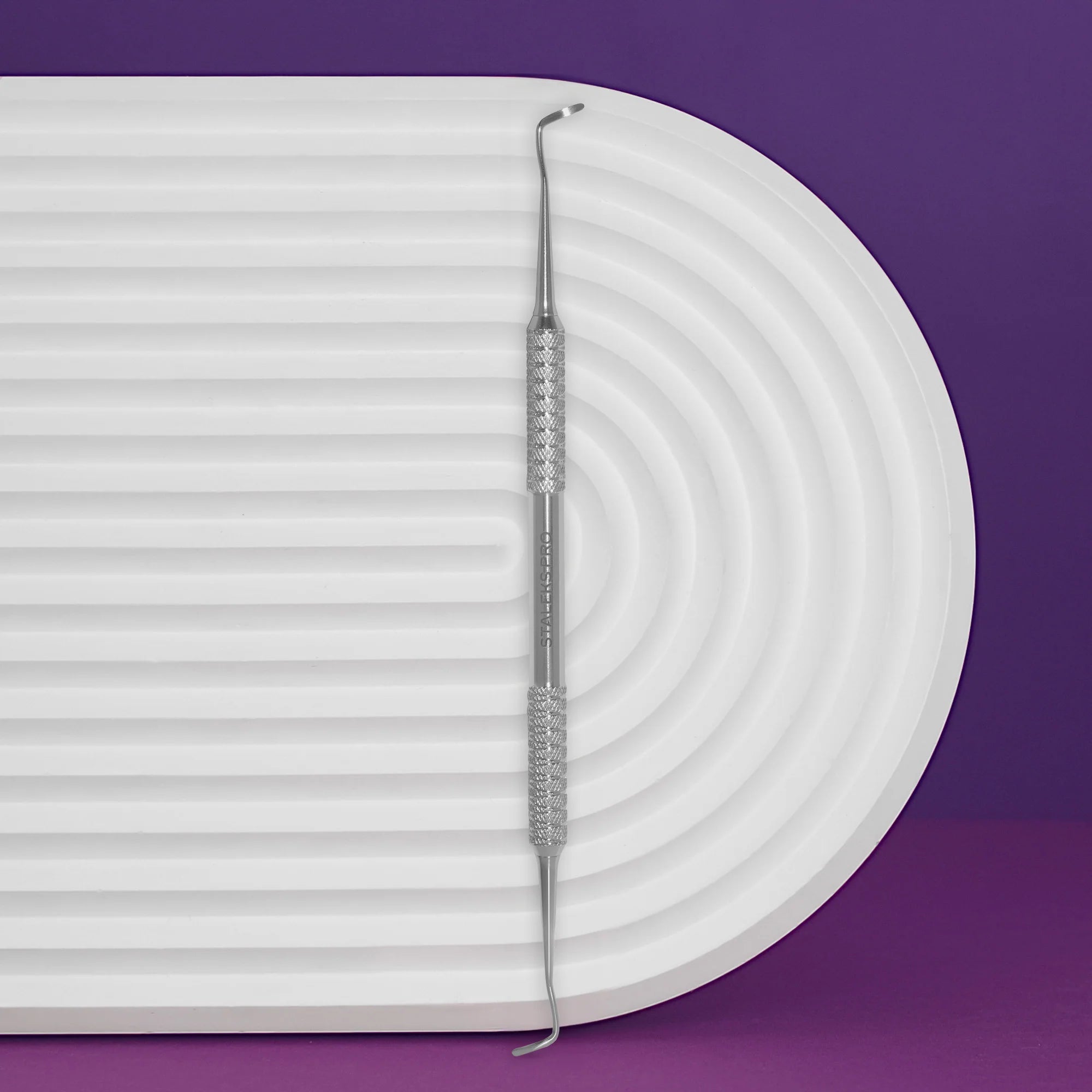 A double-ended pedicure curette with a white handle, placed on a white surface with a purple background.