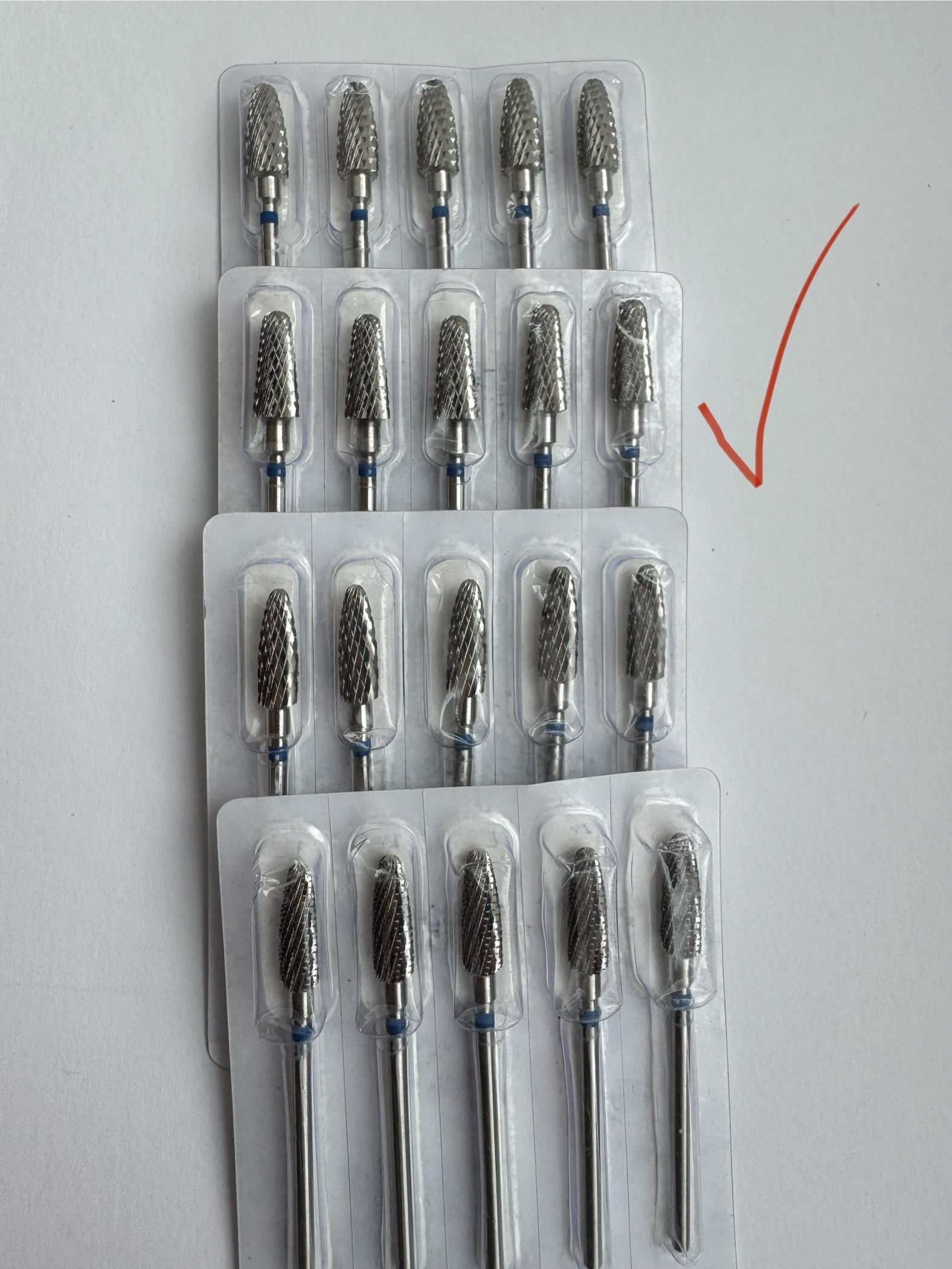 A set of carbide nail drill bits with blue rings, packaged individually in clear plastic except one bit with a red ring packaged in a different way, indicated by an arrow.