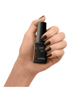 Hand holding a bottle of Kinetics nail polish with dark brown nails on a white background