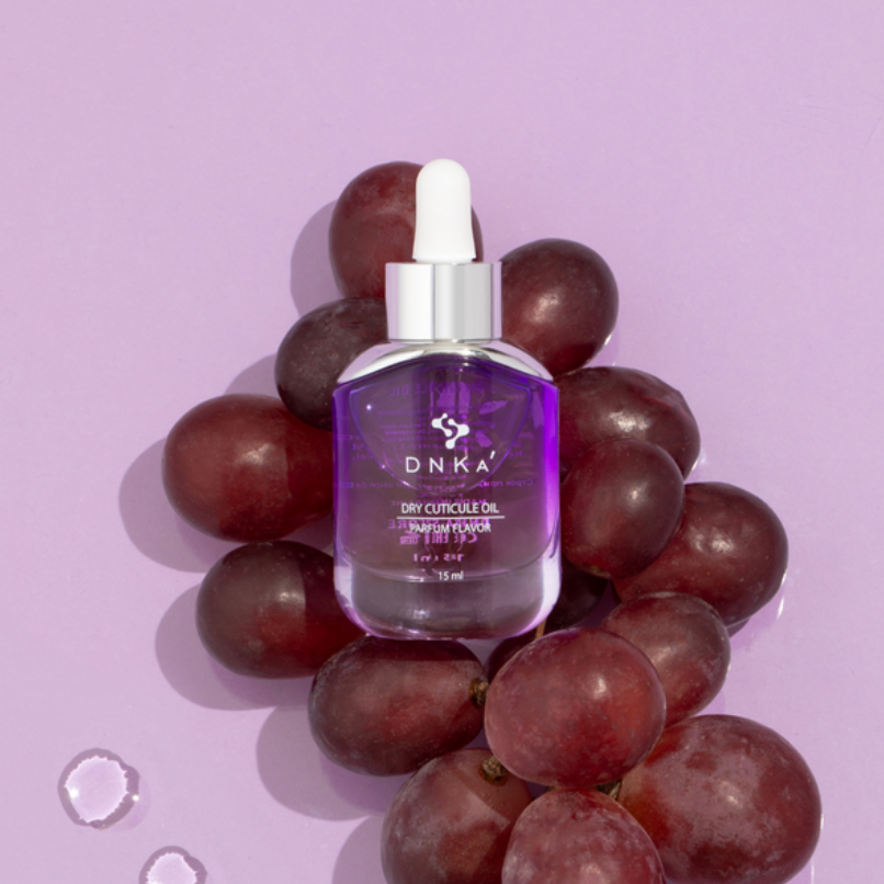 Bottle of DNKA dry cuticle oil surrounded by grapes on a light purple background