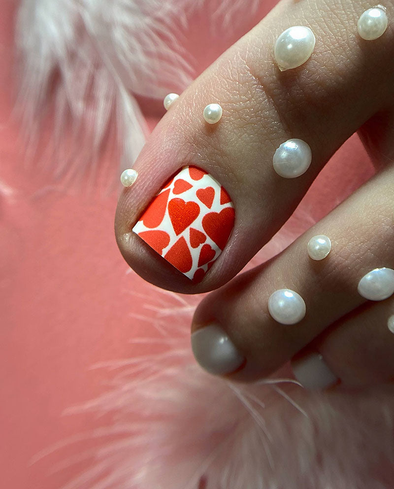 Nail art with red heart design and white pearls on a pink background