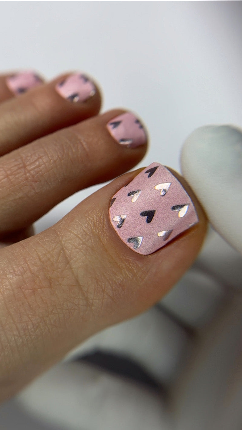 Close-up of a hand with pink nail polish featuring black heart designs on a blurred background.