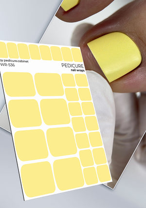 Sheet of yellow pedicure nail wraps with a person wearing a yellow nail wrap on their finger.