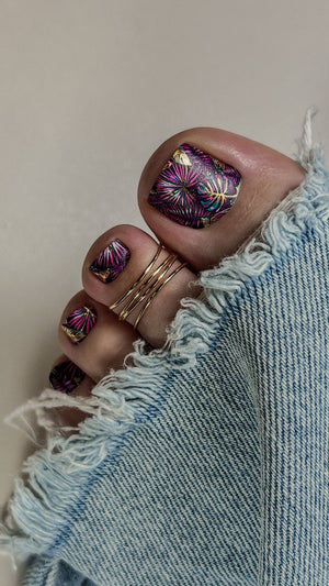 Close-up of toes with purple floral nail design wearing gold rings, against a denim background.