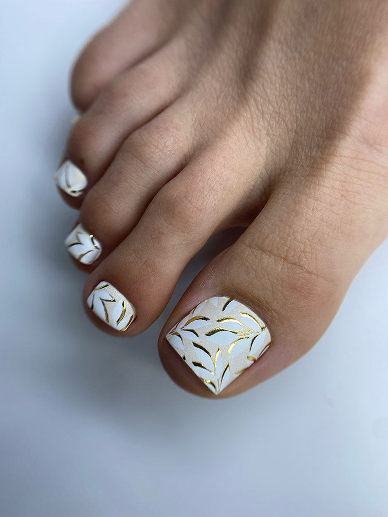 Toe nails with white nail polish and gold patterns on a light background