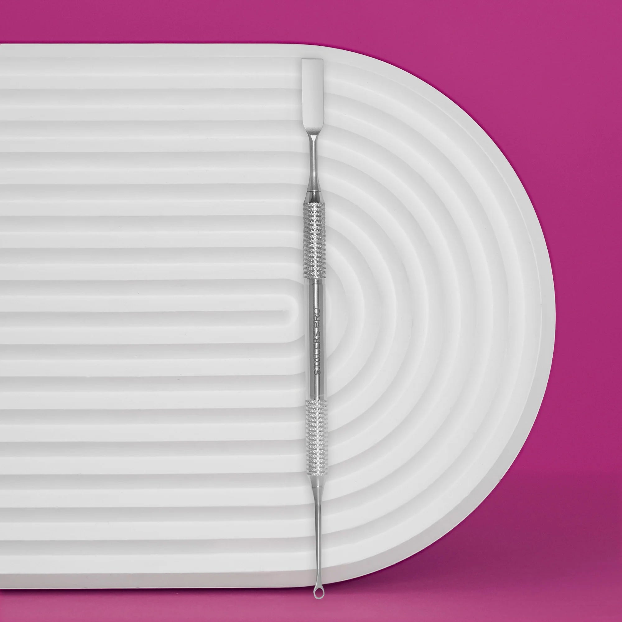A stainless steel manicure pusher with a corrugated handle, placed on a white ribbed surface with a pink background.