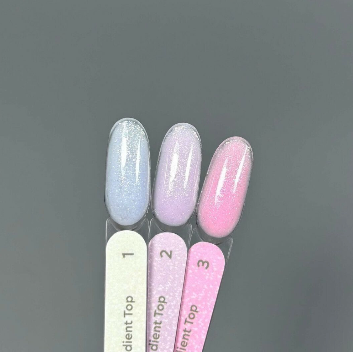 Three nail polish color swatches on nail tips labeled 1, 2, and 3, showing a light icy blue glitter shade, a pale lavender glitter shade, and a soft pink polish, with pink and lavender nail files below.