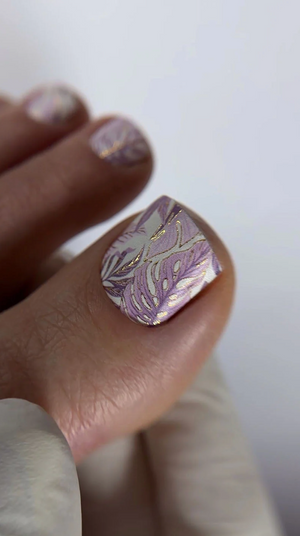 Close-up of a hand with a nail featuring a purple and silver leaf design on a plain background.