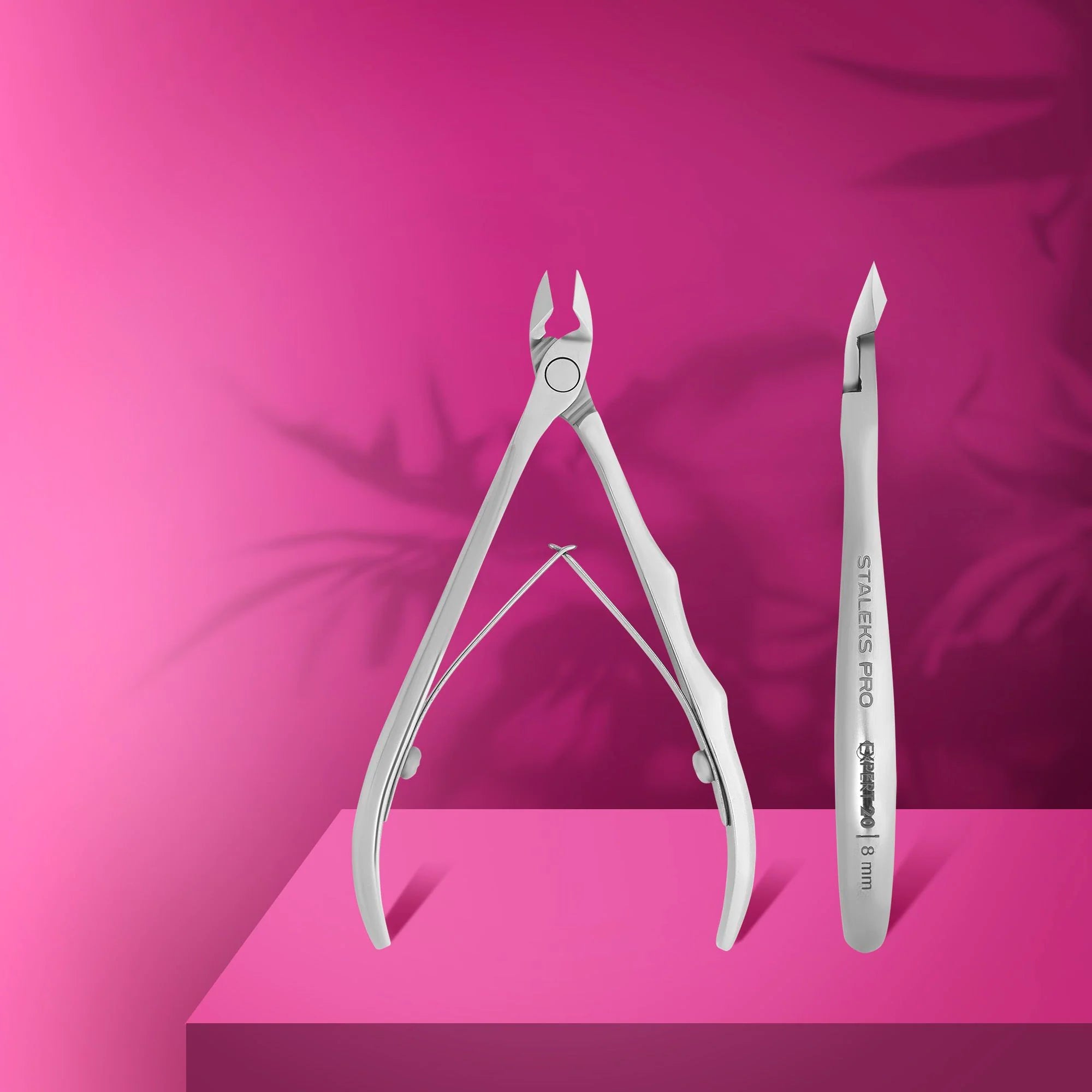 A pair of professional stainless steel cuticle nippers with straight cutting edges and a 'heel' protrusion, displayed on a pink platform with a plant in the background.