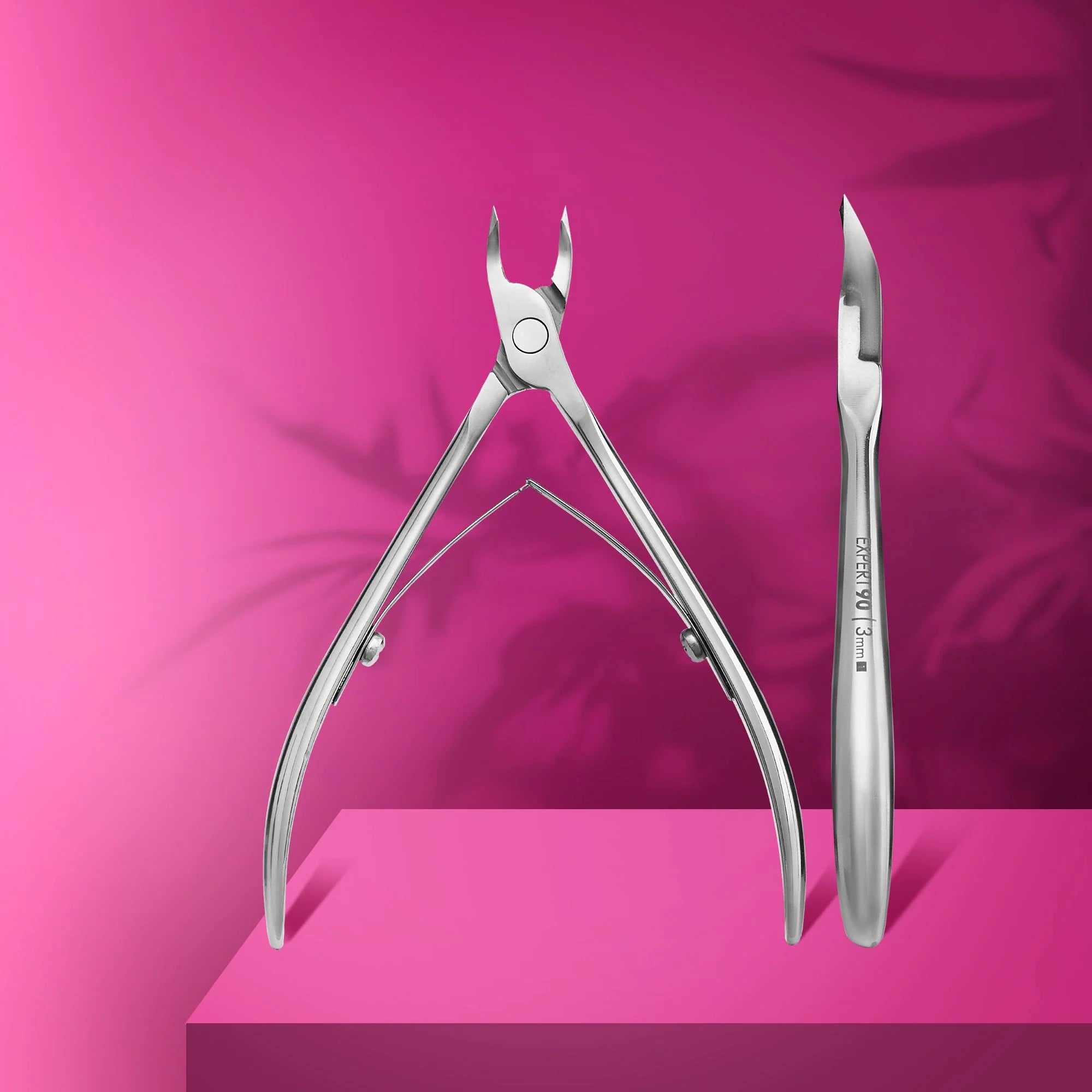 A pair of professional cuticle nippers with straight cutting edges and elongated handles, displayed on a pink platform with a blurred pink background.