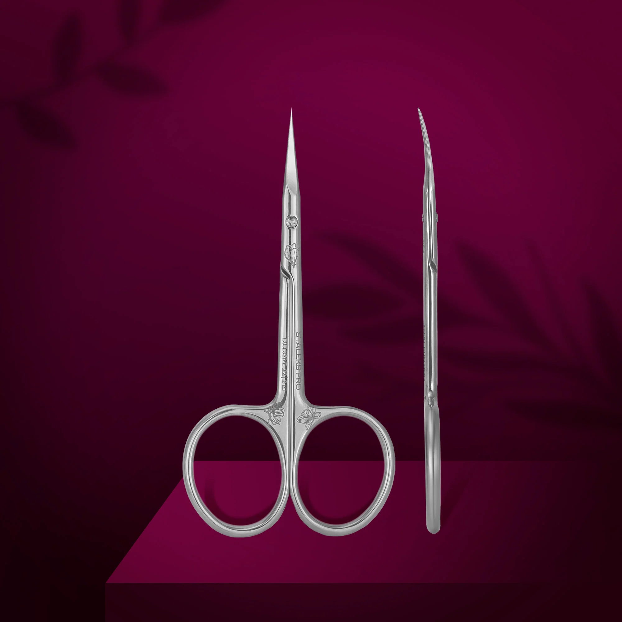 A pair of professional cuticle scissors with curved blades, displayed on a purple background with a magnolia leaf motif.
