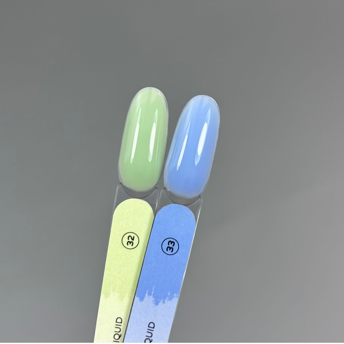 Two artificial nail tips in glossy pastel shades, one light green and one light blue, mounted on matching green and blue nail files against a smooth gray background.