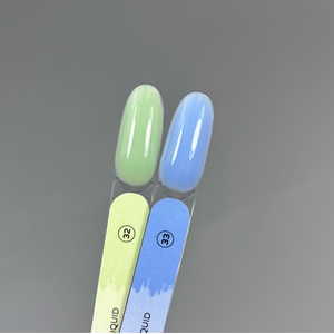 Two artificial nail tips in glossy pastel shades, one light green and one light blue, mounted on matching green and blue nail files against a smooth gray background.