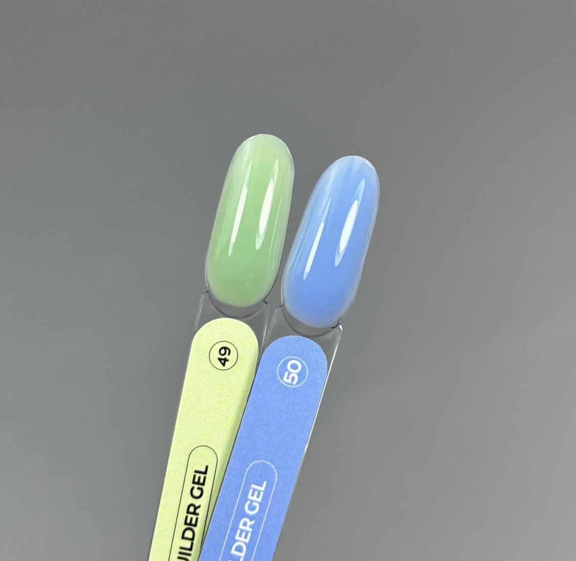 Two nail polish swatch tips on a clear plastic display stand, one in light mint green and one in light sky blue, with the text 'WILDER GEL' visible on the stands against a smooth gray background.